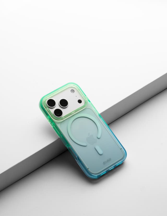 CLICK Clear Ombre MagSafe case in a green-to-blue gradient, tilted against a gray ledge on a white backdrop